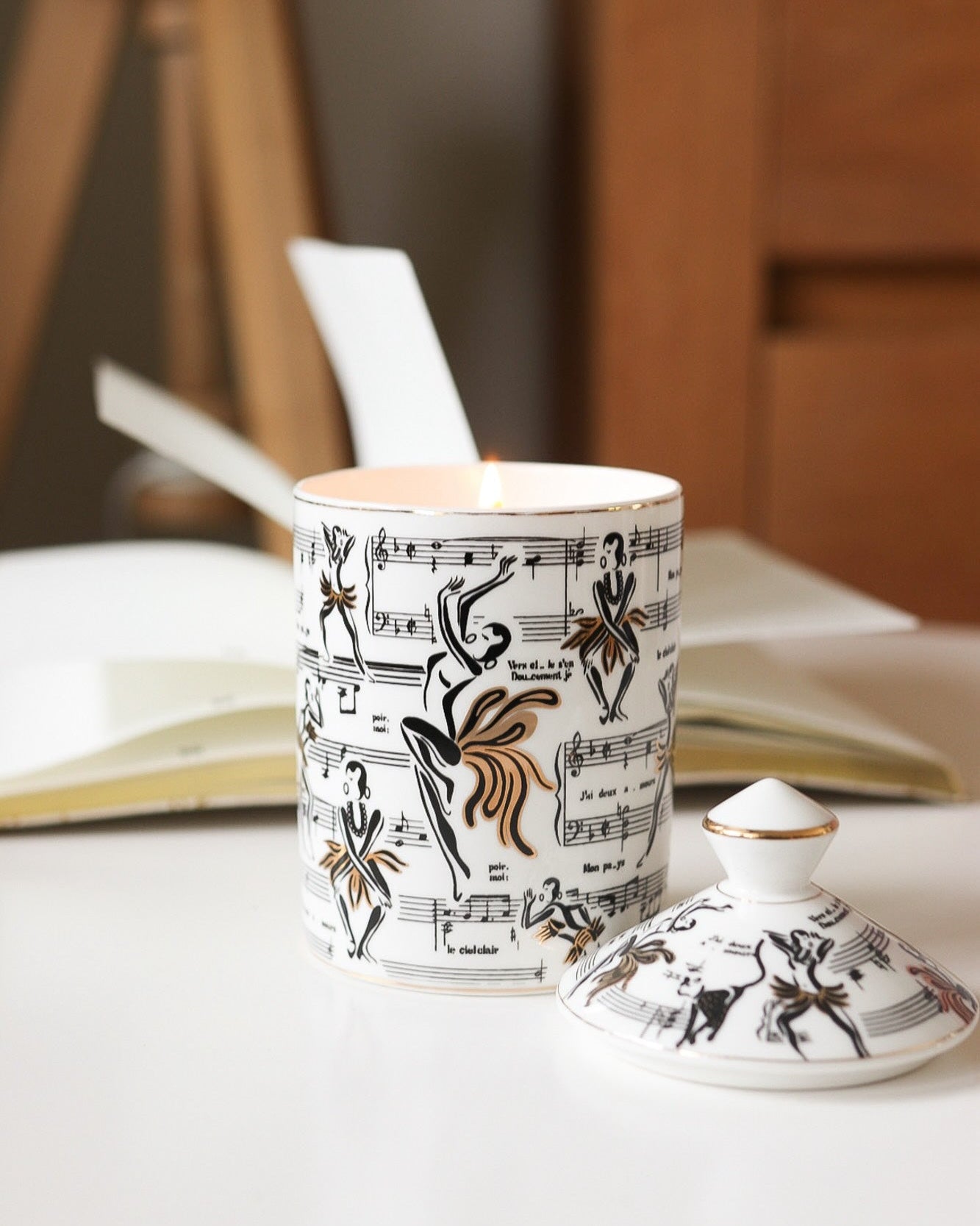 This is a lifestyle image of our Josephine ceramic candle with the lid next to the candle featuring our original illustrations of Josephine Baker dancing along with the sheet music of the song J'ai deux amours.