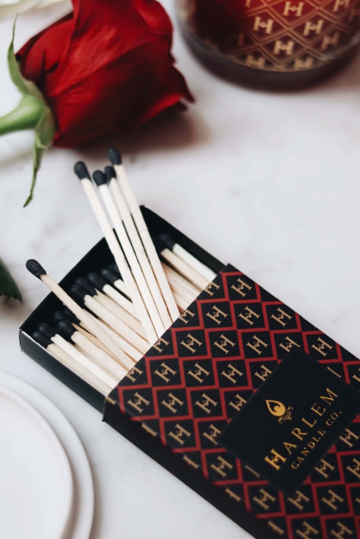 Red, black and gold Match box containing 3 inch matches with black tips. The box has the Harlem H pattern in an art deco style printed on the box. The image is on next to flowers.