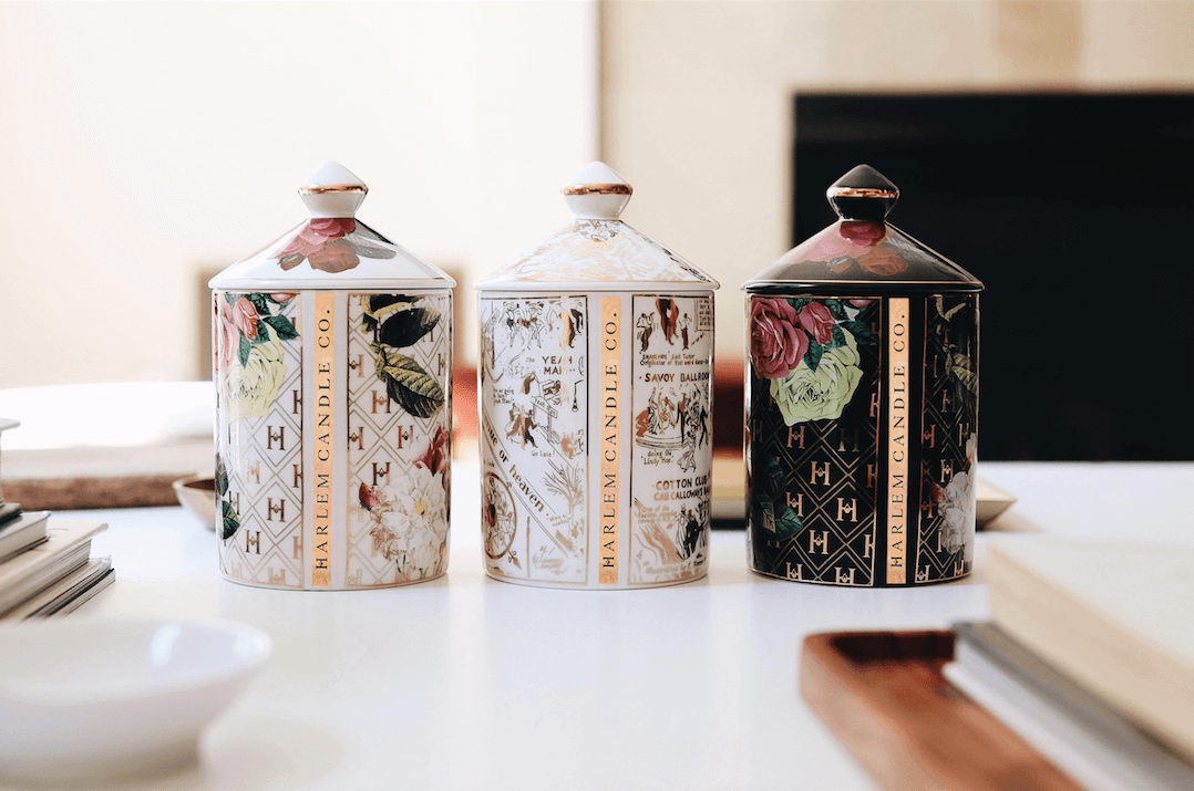 All three of our Ceramic Gold Collection candles: the “Love” by James Baldwin Ceramic Luxury Candle, the “Lady Day” White Floral Ceramic Luxury Candle, and the “Speakeasy” Harlem Map Ceramic Luxury Candle. A lifestyle image on a white table with books and wooden trays.