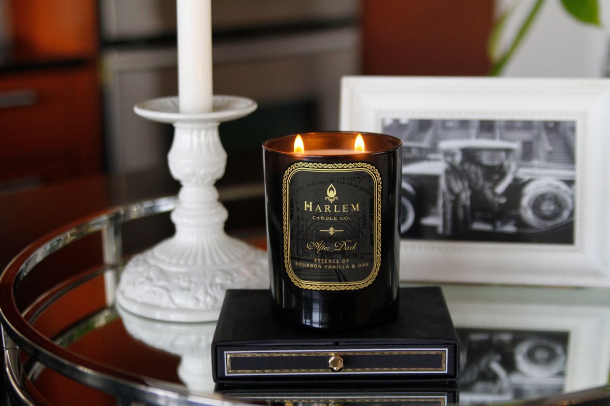 This is a Lifestyle image of the black and gold After Dark candle in a beautiful setting with home accents surrounding the candle and a vintage black and white image in the background.