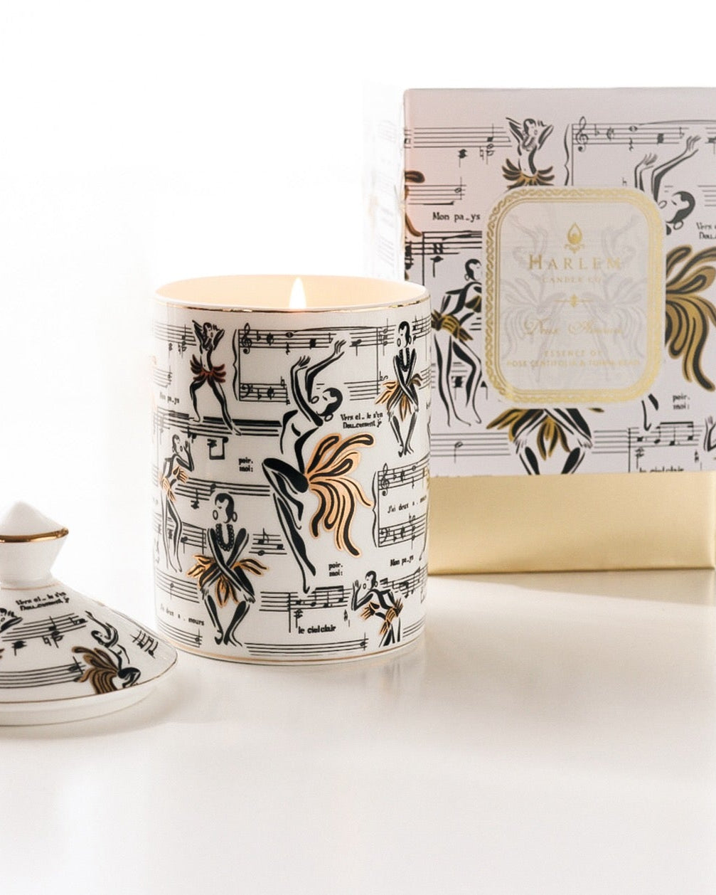 This is a lifestyle image of our Josephine ceramic candle with the lid next to the candle featuring our original illustrations of Josephine Baker dancing along with the sheet music of the song J'ai deux amours.  The candle in this image is featured next to the decorative box.