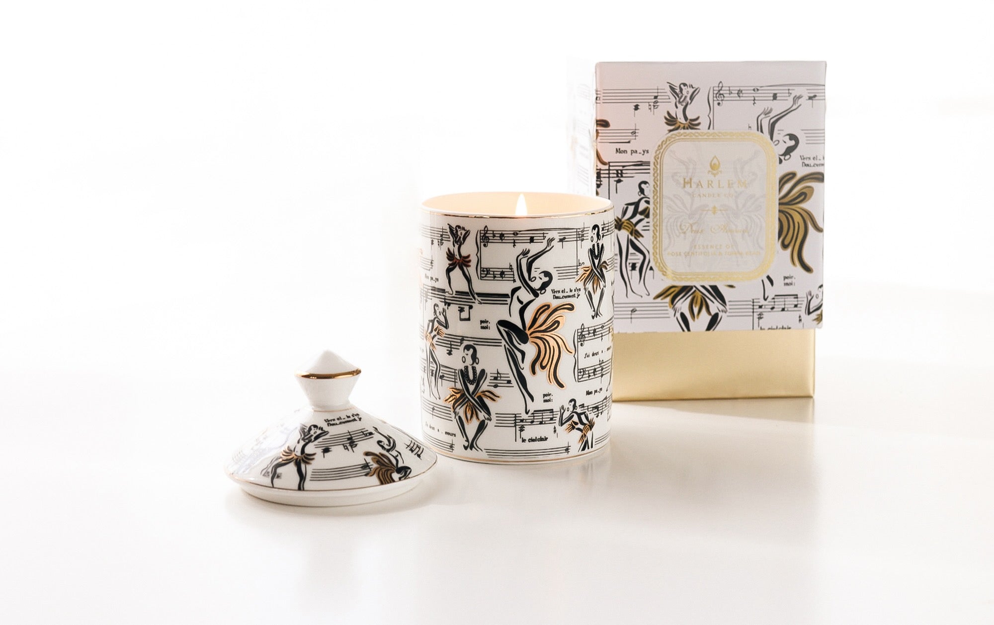 This is a lifestyle image of our Josephine ceramic candle with the lid next to the candle featuring our original illustrations of Josephine Baker dancing along with the sheet music of the song J'ai deux amours.  The candle in this image is featured next to the decorative box.