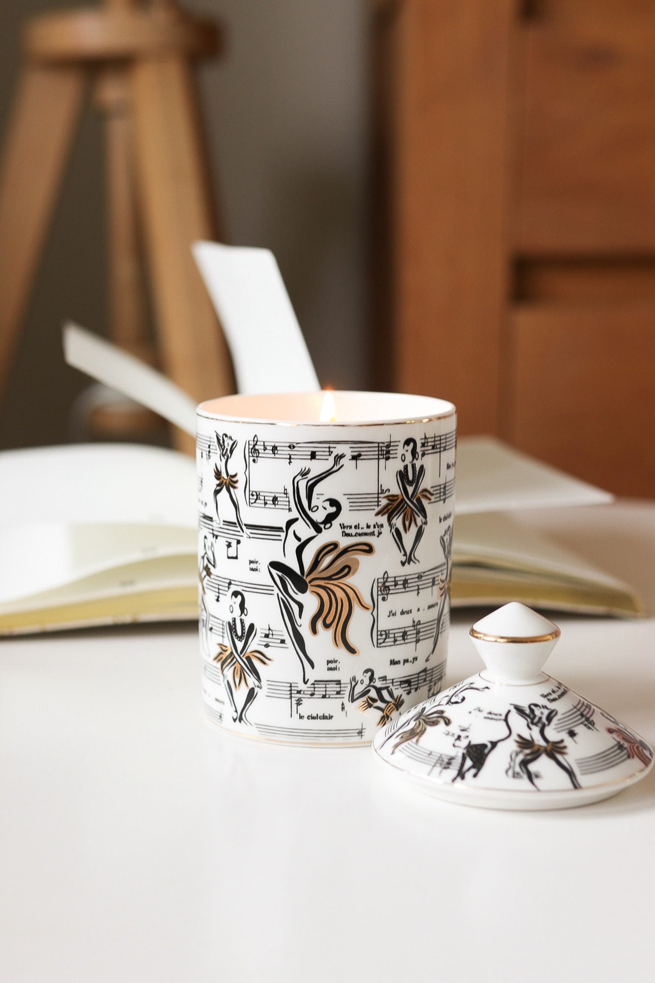 This is a lifestyle image of our Josephine ceramic candle with the lid next to the candle featuring our original illustrations of Josephine Baker dancing along with the sheet music of the song J'ai deux amours.