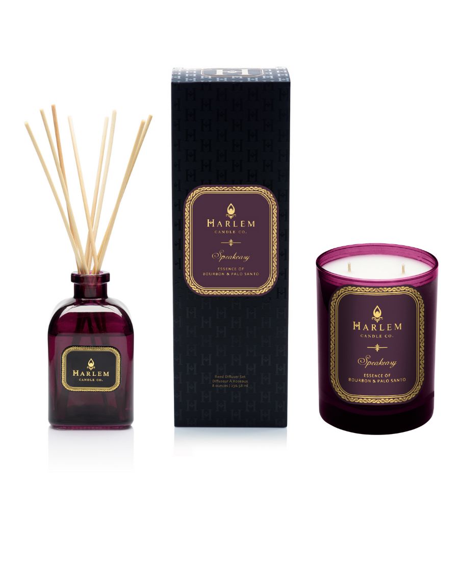 Our 8 fluid oz. Speakeasy Reed Diffuser with reeds,and 11oz Speakeasy Candlein a purple glass vessel sitting next to its decorative box on a white background.