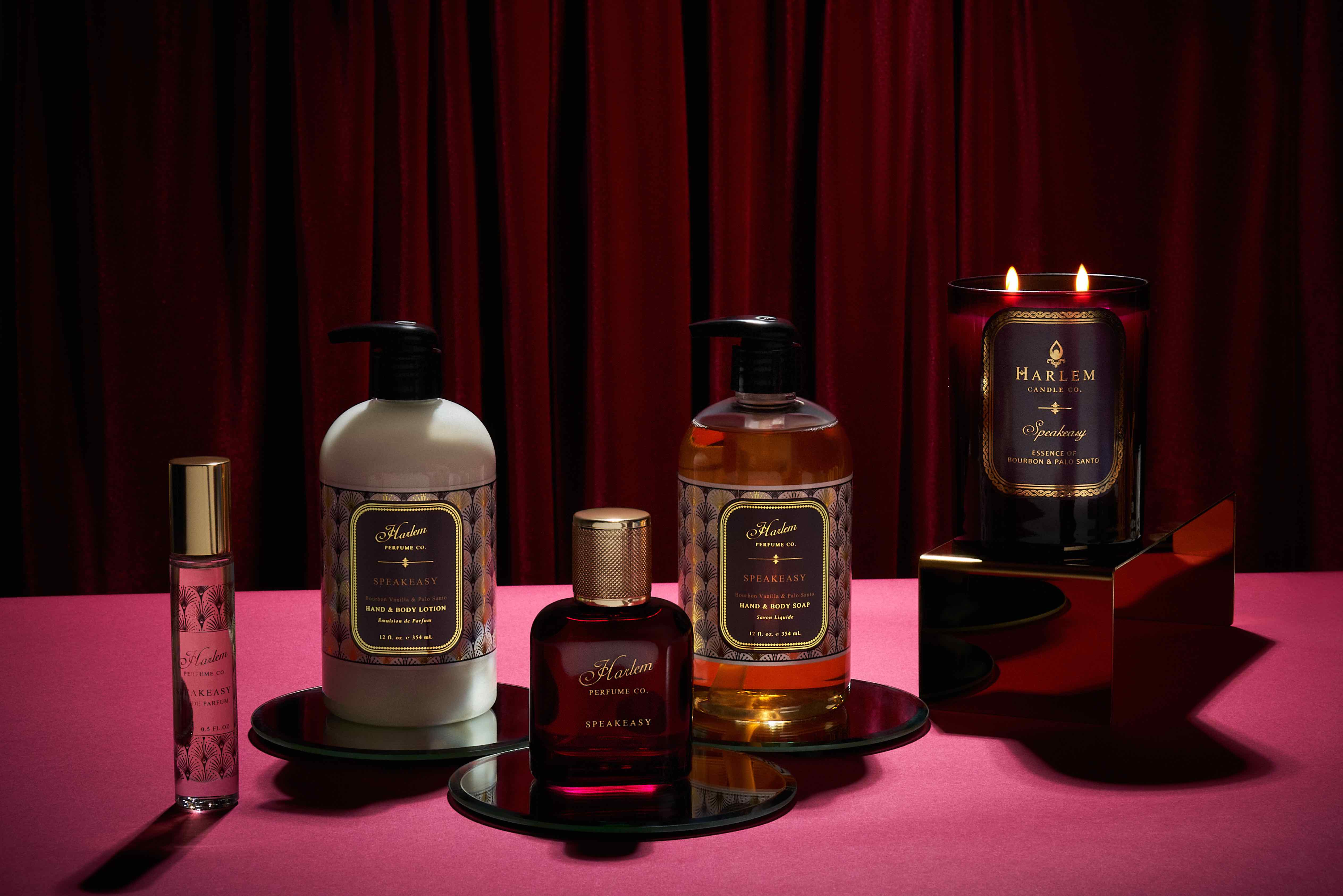 This is an image of our full speakeasy, perfume soap, lotion, and candle collection. The products are featured on a red table surface with red velvet curtains in the background.