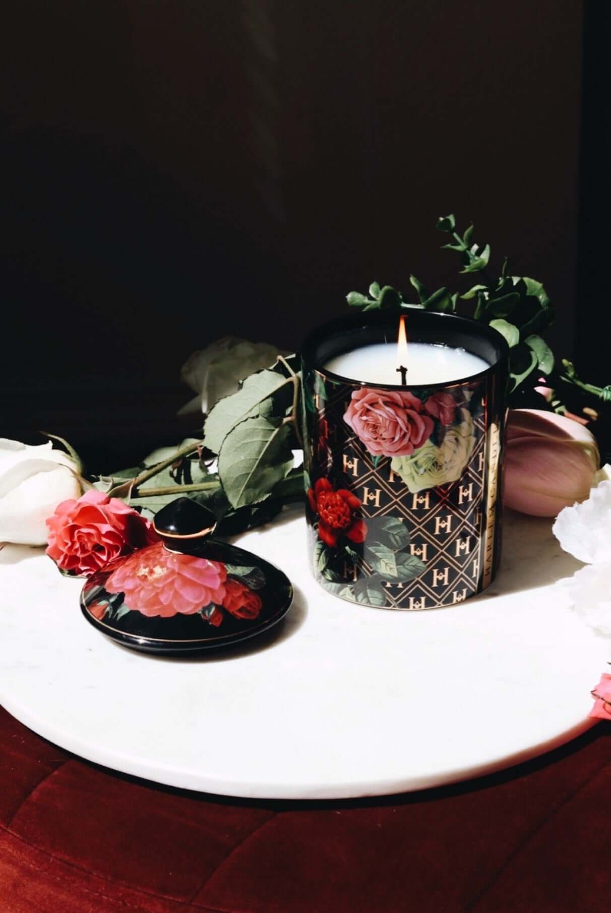 This lifestyle image features our lit Love James Baldwin ceramic candle with the lid next to the candle, on a white background with flowers and greens.