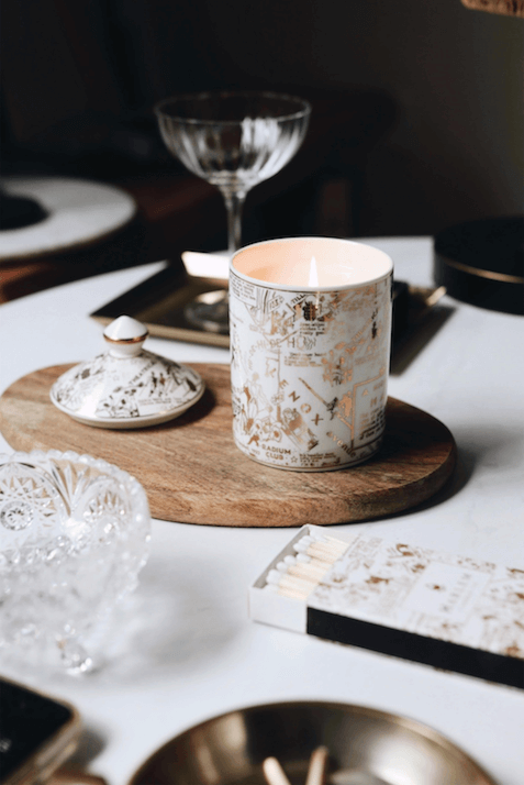 A lifestyle image of our 1 wick Our Speakeasy Map Ceramic Candle with Lid with the Night Club Map of Harlem etched in gold on the ceramic placed on a wooden coaster at a set dinner table, lit, with the ceramic top sitting next to the candle.