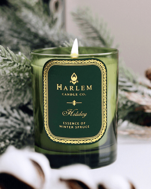 A lifestyle image of our Holiday 11 oz 1 wick candle, lit, sitting on a side table with beautiful winter greens surrounding the green candle.